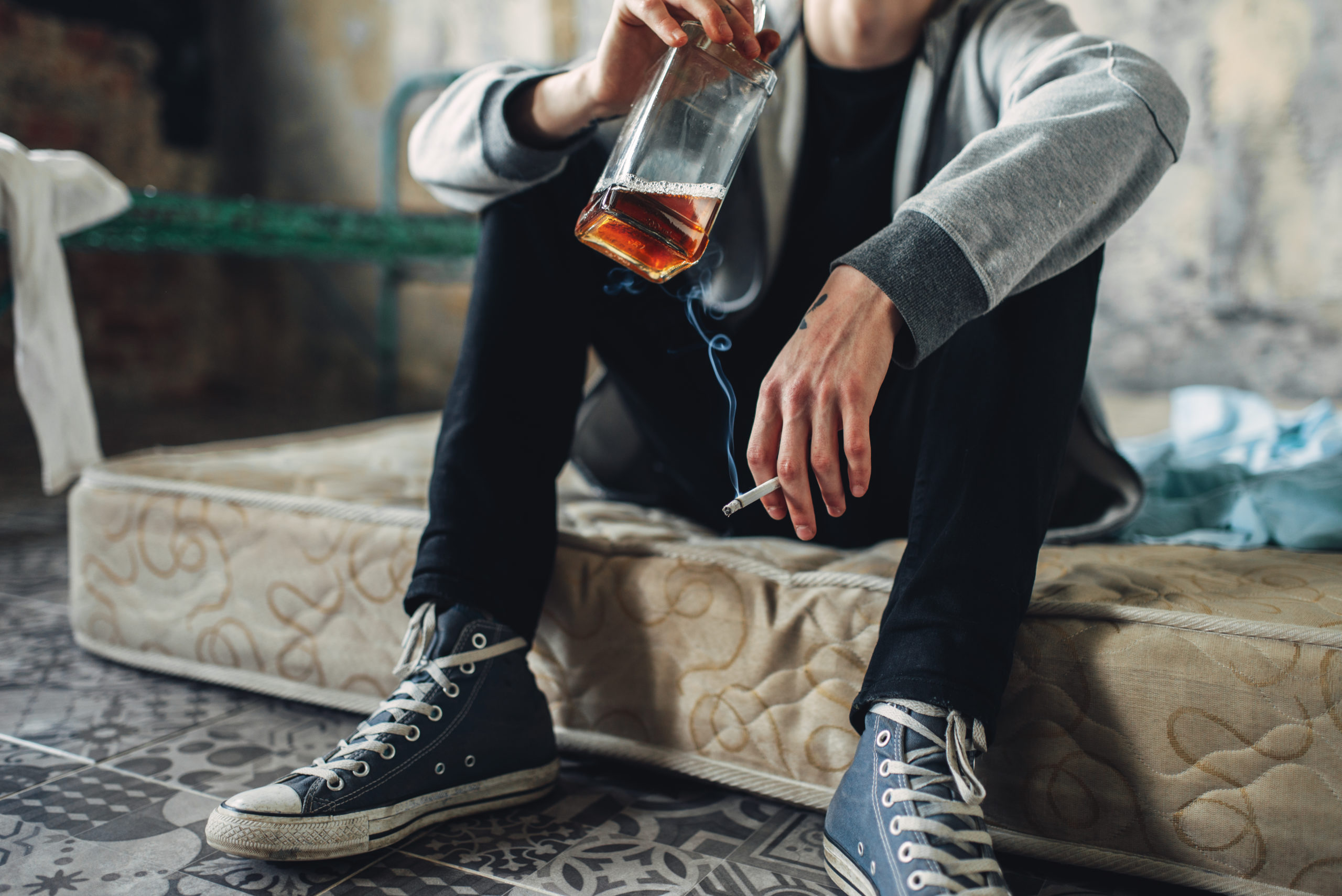Addicted male person sitting on the mattress with cigarette and bottle of alcohol in hands, grunge room interior on background. Addiction concept