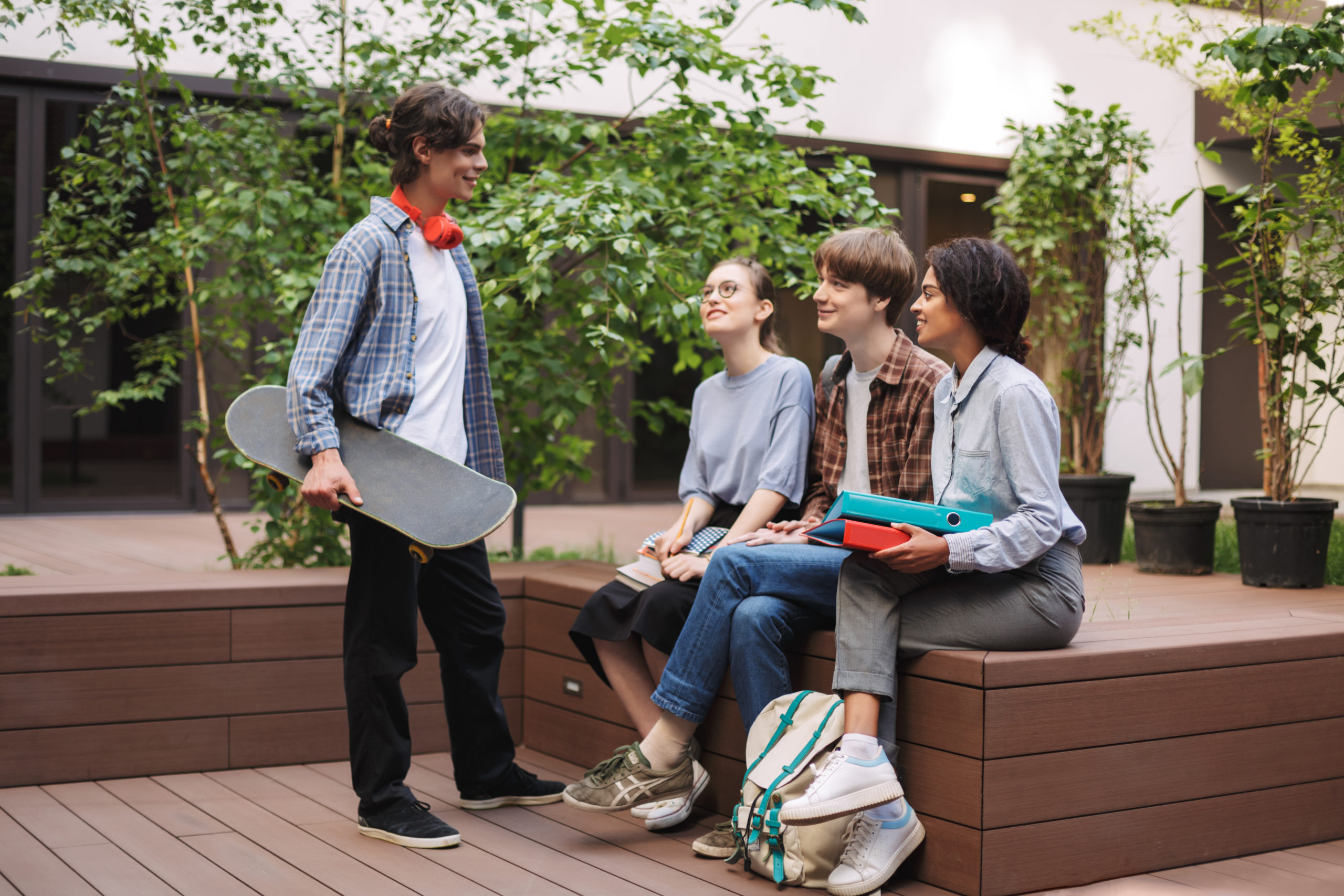 Group of young cool students sitting on bench and speaking while spending time together in courtyard of university